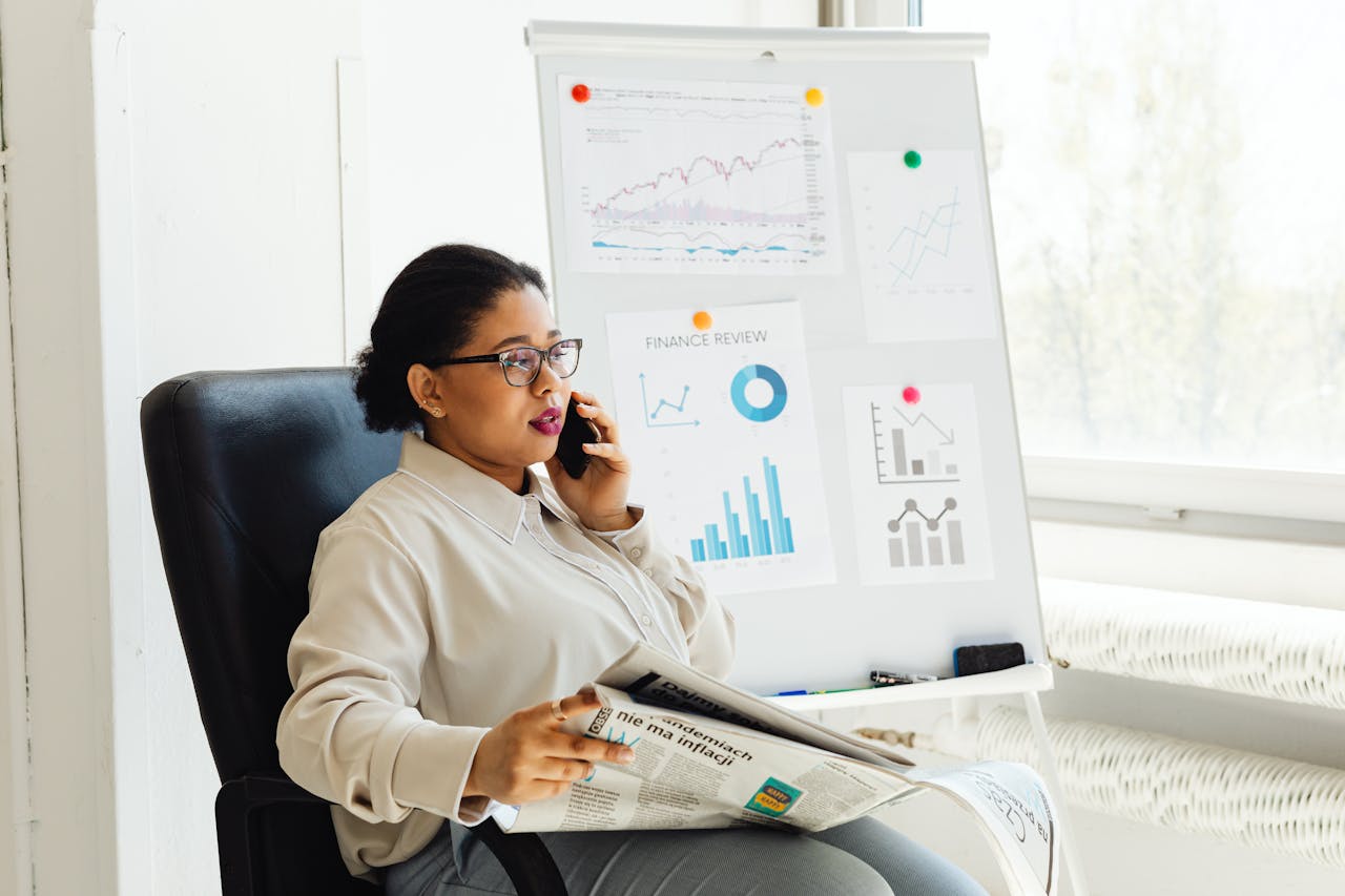 brand-01 Businesswoman multitasking with phone and newspaper in office, examining financial charts.